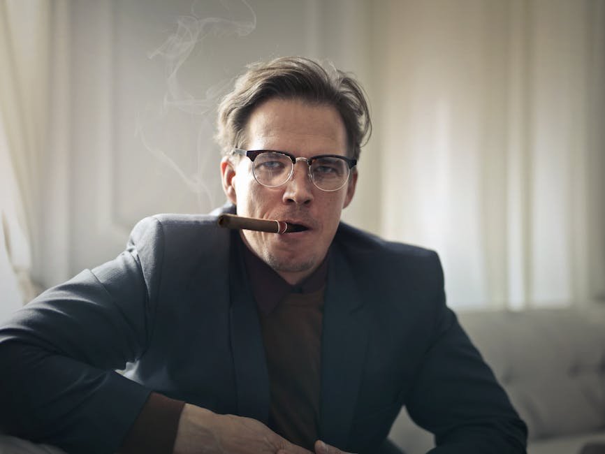 A stylish man in glasses smokes a cigar indoors, exuding confidence and sophistication.