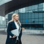 Confident businesswoman walking outside modern office building holding laptop and coffee.